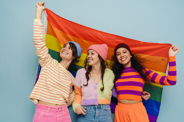 Wall Mural - Three joyful girls holding lgbt flag and hugging while standing isolated over blue background