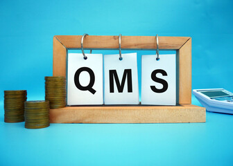 QMS. wooden cubes. Writing on paper hangs on a wooden block. stack with coins. Selective focus on QMS. short for Quality Management System.