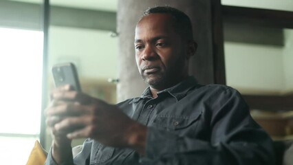Portrait of confident mature african american man hold smartphone scrolling social media texting browsing online checking email and looking at the camera while sitting on couch at home