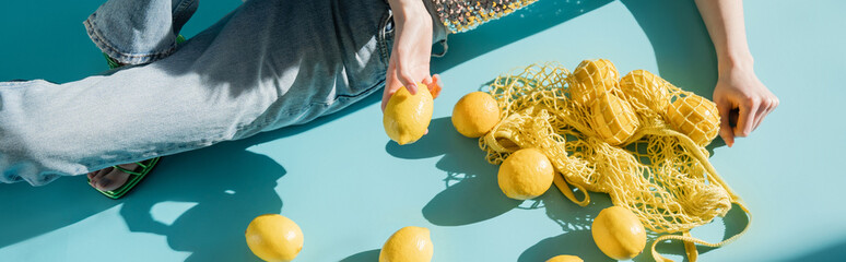 Wall Mural - high angle view of young woman in shiny top with sequins and jeans sitting near string bag and ripe lemons on blue, banner.