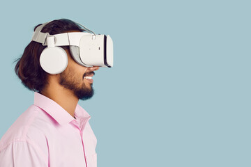 Studio shot of an ethnic man in a VR headset enjoying a virtual reality experience. Profile view of a happy Indian man wearing new VR glasses standing isolated on a light blue copy space background