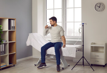 Young man sitting on a medical bed at a modern clinic or hospital, drinking a glass of water and receiving intravenous anti stress vitamin therapy treatment through an IV drip line infusion system