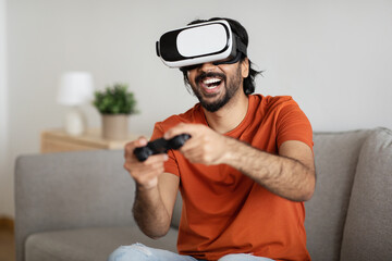 Cheerful young middle eastern guy in vr glasses and casual plays online game on computer with joystick