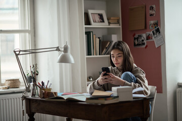 Young teenage girl studying and scrolling smartphone in her room.