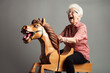 Funny old woman riding a wooden toy horse and laughing loudly. Generative AI