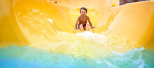 Cheerful Blue-eyed Kid On A Water Slide In The Water Park, A Little Boy Merrily Slides Into The Water Along The Slide In The Amusement Park, Rest With Children In The Water Park
