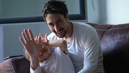 Father and child waving hello to camera sitting on sofa indoors. Dad and little boy saying HI with hand. Parenting lifestyle concept