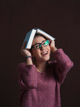 Portrait Of Teenage Girl Wearing Eyeglasses, Holding Open Book Over Head With Anxious Expression, Studio Shot On Black Background