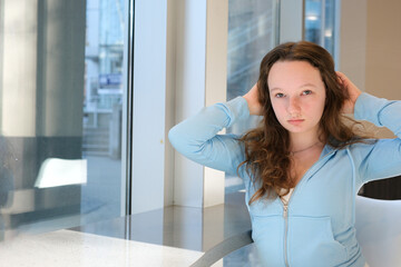 beautiful young girl teen at table by window straightens hair looks into frames sports blue light jacket brown hair asian eyes white skin puffy lips european ukrainian young woman growing up