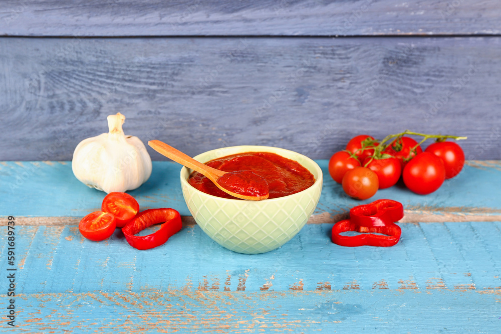 Bowl with tasty tomato paste and fresh vegetables on blue wooden background