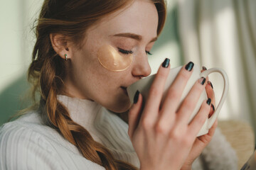 Wall Mural - close-up portrait ginger lady sitting on sofa and drinking coffee while wearing eye patches. natural