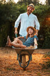 © _KUBE_ - Happy bearded man in a straw hat is pushing a cart with a cheerful little girl. Father and daughter enjoy summer together in the backyard. Vertical. The concept of gardening