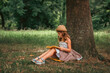 © _KUBE_ - School holidays. Pretty schoolgirl girl in a straw hat and dress is sitting on the ground by a tree and reading a book. The concept of independent extracurricular children's education