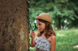 © _KUBE_ - Children's education. Portrait of curious little girl in a straw hat looks at the tree bark through a magnifying glass. Copy space. The concept of scouting and childhood