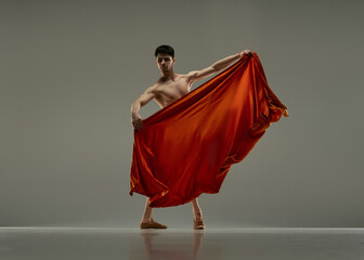 Young handsome man, ballet dancer making performance with red silk fabric against grey studio background. Concept of art, classical dance, inspiration, creativity, fashion, beauty, choreography