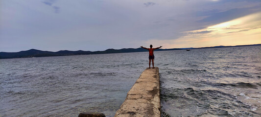 A guy with a backpack and arms outstretched stands on the pier. Sea coast in the city of Zadar. Adriatic Sea. Dalmatia. Croatia. Europe