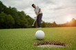 © somchai - White golf ball rolling down golf hole on putting green with evening golf course.
