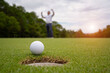 © somchai - White golf ball rolling down golf hole on putting green with evening golf course backdrop.
