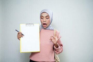 Smiling Asian Muslim female student in pink sweater with bag, holding board and pointing at empty board isolated on white background. back to school concept