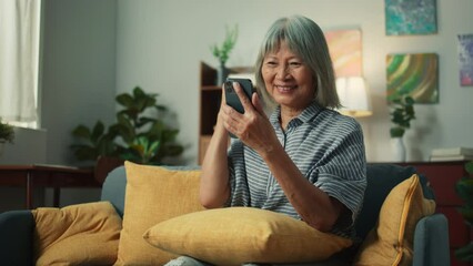 Wall Mural - Happy Asian elderly woman smile holding mobile phone checking email received good news, feeling surprised excited. Cheerful aged female looking at smartphone screen sit on sofa in home living room.