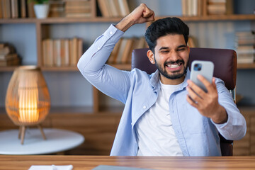 Young successful businessman inside the office holds a smartphone in his hands, the man holds his hand up a gesture of success and triumph, celebrating victory.