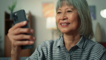 Wall Mural - Happy attractive Asian elderly woman using smartphone video conference call in cozy living room. Happy old aged female using mobile phone technology chatting talking with family or friends at home