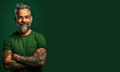 © piai - Smiling bearded tattooed mature man in green t-shirt with crossed arms stands against the green background. Generative AI