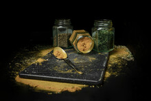 Three Kinds Of Herbs Or Spices In Glass Jars Sitting In A Row On A Black Background With A Spoon And Dustings Of Herbs And Spices On The Countertop; Studio