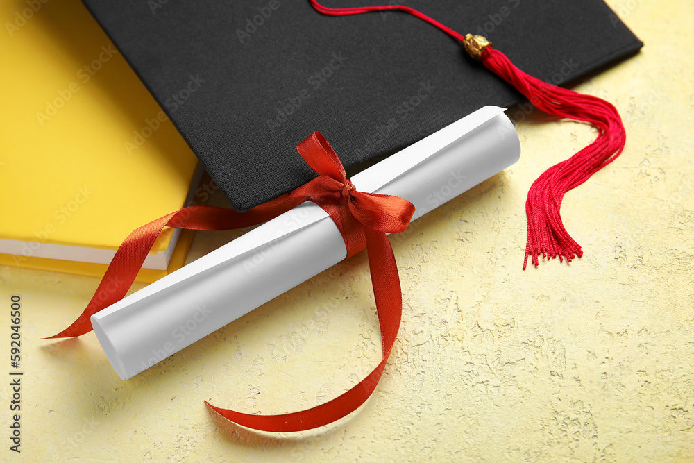 Diploma with red ribbon, graduation hat and book on yellow table