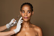 Beautiful black middle aged woman getting facial injection in cheekbones zone, standing over brown studio background