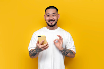 Wall Mural - unshaven asian man in white t-shirt holds phone and shows with his hand ok on yellow isolated background