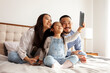 © Богдан Маліцький - asian family sit on the bed and communicate via video call, little korean girl with mom and dad takes selfie