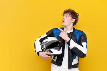 young guy motorcyclist in leather jacket holds helmet and points to the copy space on yellow isolated background