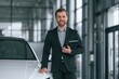 © standret - With tablet in hands. Man in black suit is indoors in the car dealership near automobile