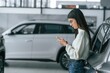 © standret - Young woman is leaning on the car, holding smartphone. Other automobiles are indoors