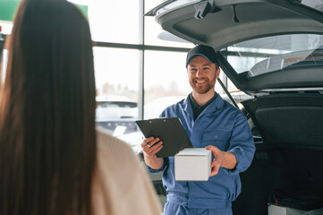 Man is holding box and notepad. Young woman is visiting auto mechanic in garage. Repair service