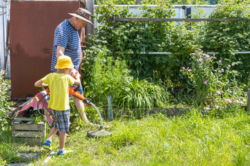 A man in a hat and a child with an electric lawn mower mowing the lawn. Grandfather and grandson together