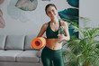 © ViDi Studio - Young fun strong sporty athletic fitness trainer instructor woman wearing green tracksuit hold in hand yoga mat drink water training do exercises at home gym indoor. Workout sport motivation concept.