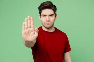 Wall Mural - Young strict serious sad brunet caucasian man he wearing red t-shirt casual clothes showing stop gesture with palm isolated on plain pastel light green background studio portrait. Lifestyle concept.