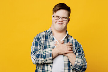 Young smiling thankful man with down syndrome wearing glasses casual clothes look camera put folded hands on heart isolated on pastel plain yellow color background. Genetic disease world day concept.