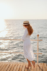 Wall Mural - A young girl in a white dress and long hair, in sunglasses and a straw hat, stands on the pier against the backdrop of the ocean at sunset