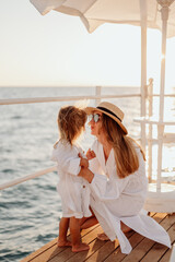 Wall Mural - A young girl, mother, kisses her little daughter. Girls in white dresses on the background of the evening ocean. Girl in a straw hat