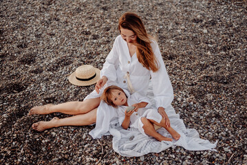 Wall Mural - A young mother and daughter in white dresses are sitting on a rocky ocean shore. Little daughter lay down on her mother and looks at the camera