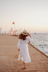 Wall Mural - A young girl with long hair in a white dress and a straw hat walks towards the ocean along a wooden pier. Hair develops the wind