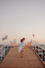 Wall Mural - A young girl with long hair in a white dress walks towards the sea along a wooden pier and holds a hat in her hands. Hair develops the wind