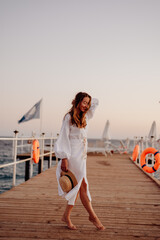 Wall Mural - A young girl with long hair in a white dress, with bare feet, stands on a wooden pier against the backdrop of the ocean at sunset and holds a hat in her hands. Wind blows hair