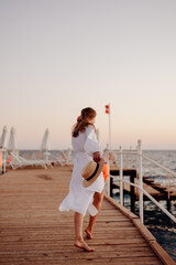 Wall Mural - A young girl with long hair in a white dress walks barefoot along a wooden pier against the backdrop of the ocean at sunset and holds a hat in her hands. Wind blows hair