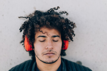 Close-up portrait of a handsome young North African man with curly hair with eyes closed, leaning against a wall while listening to music on headphones with a dreamy expression