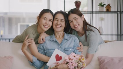 Wall Mural - Happy time Mother day grown up child looking at camera cuddle hug give flower gift box red heart card to mature mum. Love kiss care mom asia middle age adult people smile enjoy sitting at home sofa.