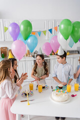 Wall Mural - happy children clapping hands and singing happy birthday song next to cake with candles and balloons.